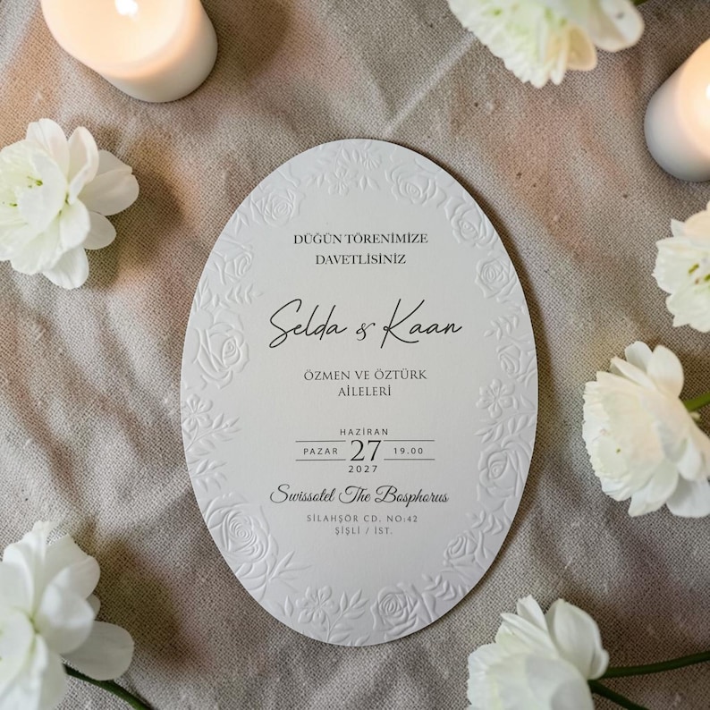 May include: An oval-shaped wedding invitation with embossed floral details and text. The invitation features the names "Selda & Kaan", event details, and the venue "Swissotel The Bosphorus". White flowers and candles surround the invitation.