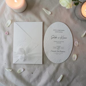 May include: White wedding invitation set with an envelope and an oval card. The envelope has embossed initials and a bow. The card features floral detailing and text. The scene includes candles and flower petals.