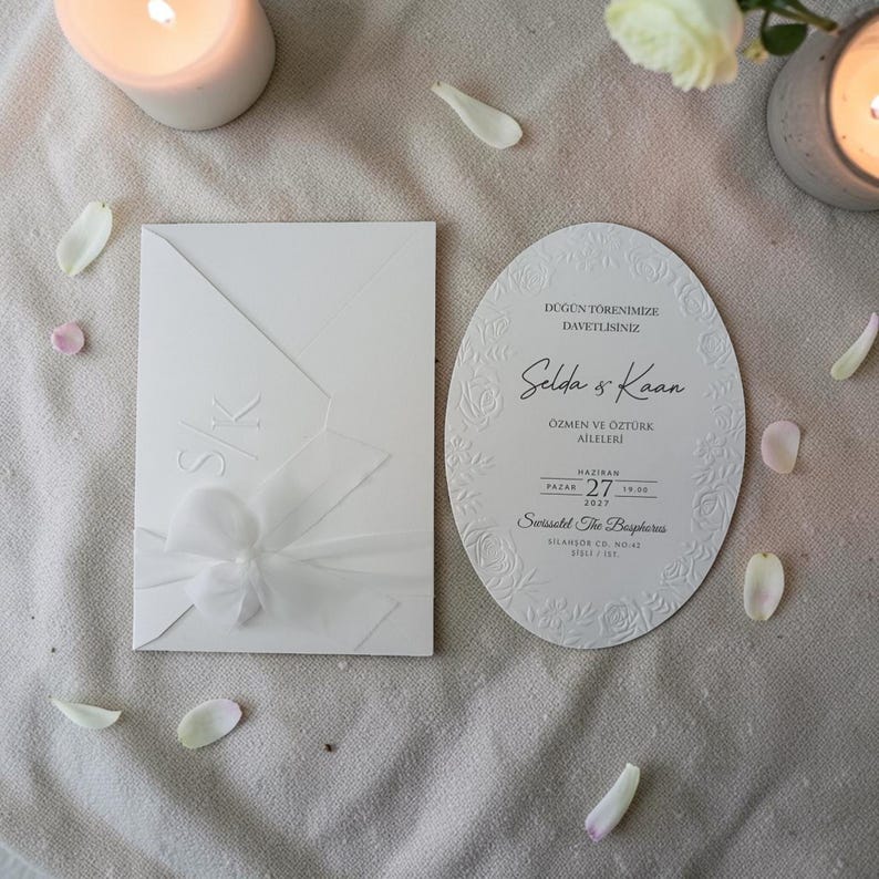 May include: White wedding invitation set with an envelope and an oval card. The envelope has embossed initials and a bow. The card features floral detailing and text. The scene includes candles and flower petals.