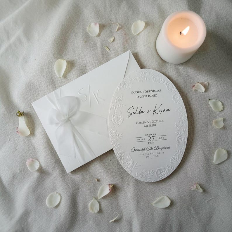 May include: A white wedding invitation set with an oval card and envelope, featuring embossed floral details and a bow. A lit candle and scattered petals add to the elegant presentation. The invitation includes the names "Selda & Kaan" and the date.