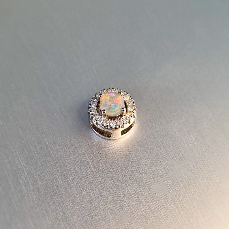 May include: A round, silver pendant featuring a central opal stone surrounded by a halo of small, clear gemstones. The opal displays a play of colors, including pink, yellow, and blue. The pendant is set against a light gray background.