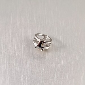 May include: A small, silver-colored charm with a geometric design. The charm has a circular shape with an open center and is adorned with small, clear stones around the bottom edge. The charm is set against a light gray background.