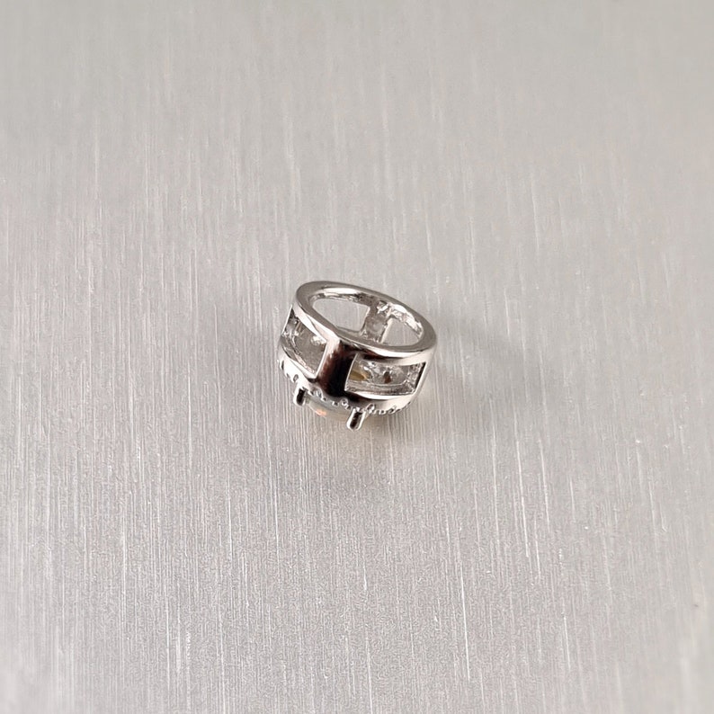 May include: A small, silver-colored charm with a geometric design. The charm has a circular shape with an open center and is adorned with small, clear stones around the bottom edge. The charm is set against a light gray background.