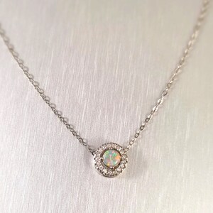 May include: A delicate silver necklace featuring a round pendant with a central opal stone surrounded by a halo of small, clear gemstones. The opal displays a play of colors. The chain is fine and simple.