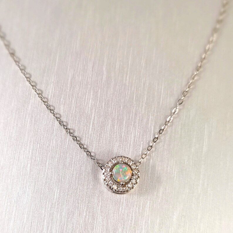 May include: A delicate silver necklace featuring a round pendant with a central opal stone surrounded by a halo of small, clear gemstones. The opal displays a play of colors. The chain is fine and simple.