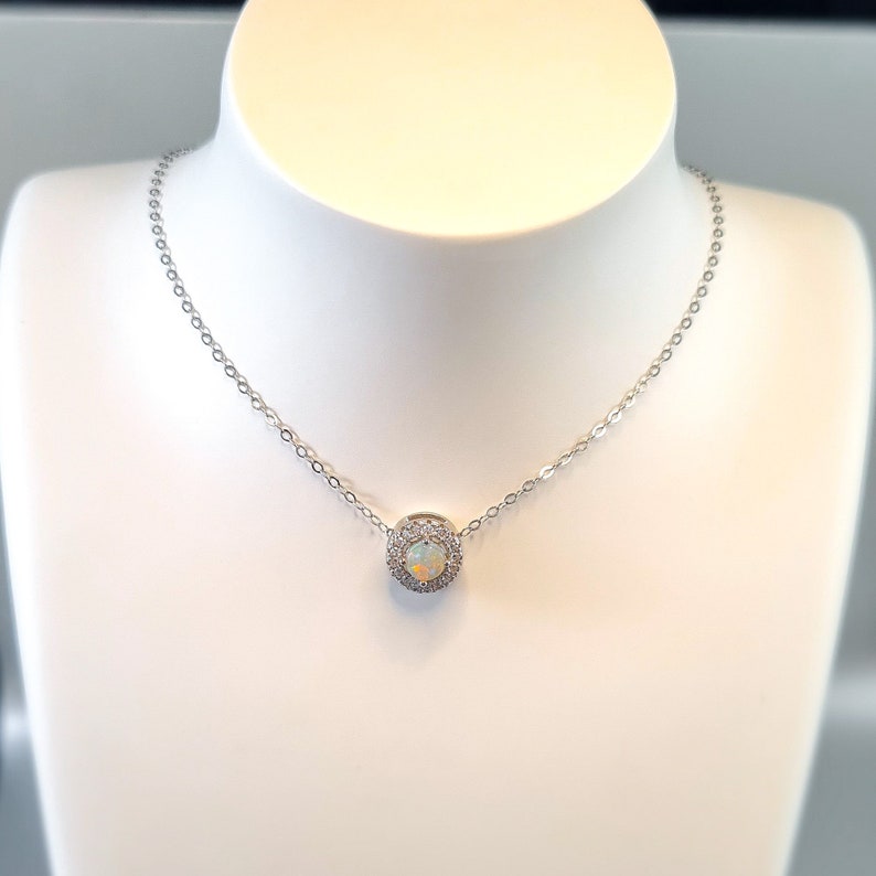 May include: A silver necklace with a pendant featuring a central opal stone surrounded by a halo of small, clear crystals. The necklace is displayed on a white mannequin bust, showcasing the delicate chain and pendant design.