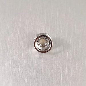 May include: A small, silver-colored jewelry finding with a circular design. The center features a light-colored, possibly pearl-like, element. The finding has a detailed, textured edge and is set against a brushed metal background.