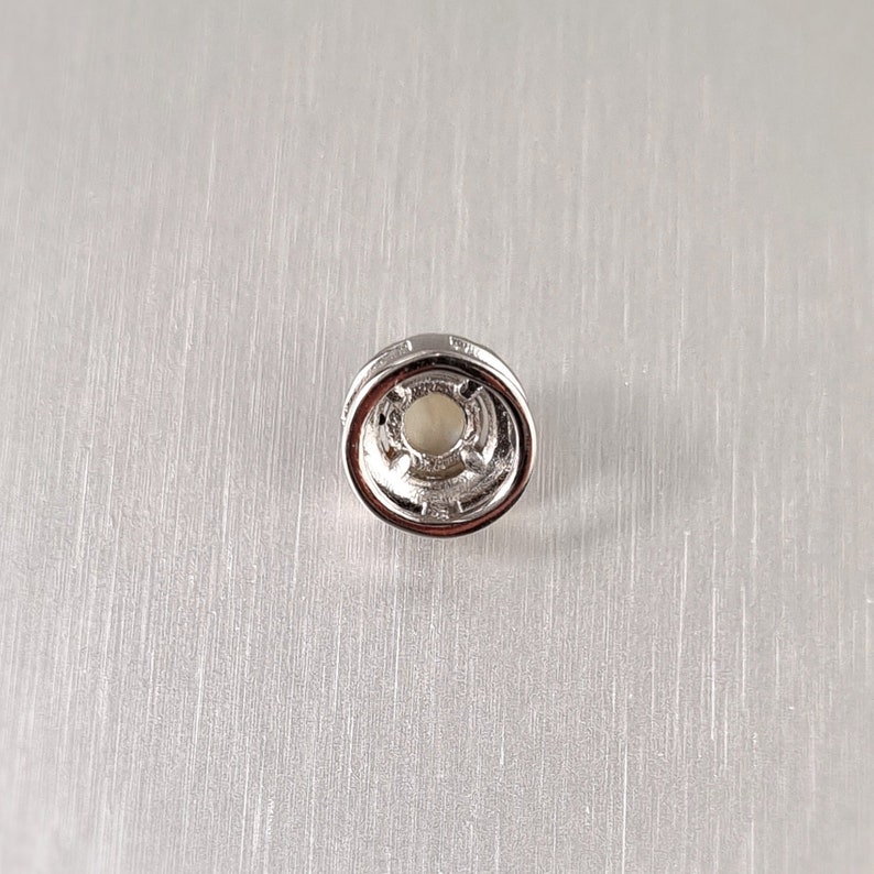 May include: A small, silver-colored jewelry finding with a circular design. The center features a light-colored, possibly pearl-like, element. The finding has a detailed, textured edge and is set against a brushed metal background.