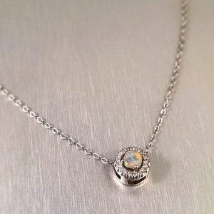 May include: A silver necklace featuring a round pendant with a central opal stone surrounded by small, clear gemstones. The opal displays a range of colors. The chain is delicate and silver in color.
