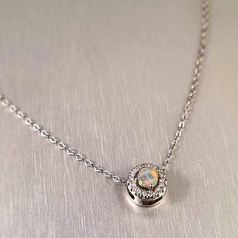 May include: A silver necklace featuring a round pendant with a central opal stone surrounded by small, clear gemstones. The opal displays a range of colors. The chain is delicate and silver in color.