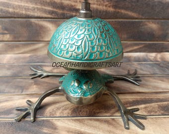 May include: A brass frog bell with a green and gold finish. The bell is shaped like a frog with a round top and a small handle. The frog is sitting on a wooden surface.