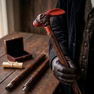 May include: A walking cane featuring a detailed, reddish-brown carved rabbit head handle with long ears, attached to a dark, textured wooden shaft with copper-colored metal accents. The cane is shown in three segments, with a small wooden box nearby.