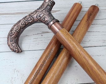 May include: A walking cane featuring a T-shaped handle with an ornate, embossed copper-toned floral pattern. The shaft is made of polished brown wood with visible grain, and the text "OCEANHANDICRAFTSART" is engraved on the side of the shaft.
