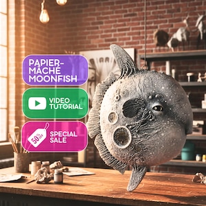 Puede incluir: Una escultura de pez luna de papel maché gris con detalles texturizados cuelga en un estudio. La escultura tiene un cuerpo redondo con cráteres lunares y una aleta. La imagen incluye texto: "PAPIER-MÂCHÉ MOONFISH", "VIDEO TUTORIAL" y "SPECIAL SALE 50% OFF".