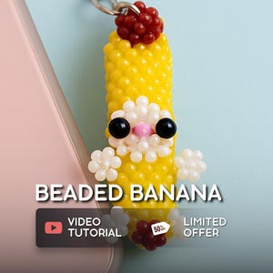 Puede incluir: Un dije de plátano con cuentas con un cierre plateado. El plátano amarillo tiene ojos de cuentas negras, detalles de cuentas blancas y una parte superior de cuentas rojas. El texto "BEADED BANANA" se muestra encima del dije.