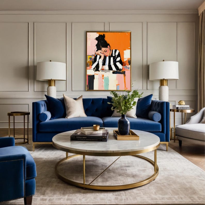 May include: A blue velvet sofa with a gold metal coffee table in a living room setting. The coffee table has a marble top and a gold metal frame. A painting of a woman in a striped shirt is hanging on the wall behind the sofa.