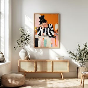 May include: A framed painting of a woman with black and white striped clothing, leaning on a table with a colorful background. The painting is hanging on a white wall above a wooden cabinet with three drawers.