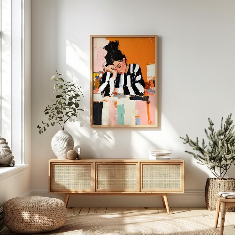 May include: A framed painting of a woman with black and white striped clothing, leaning on a table with a colorful background. The painting is hanging on a white wall above a wooden cabinet with three drawers.