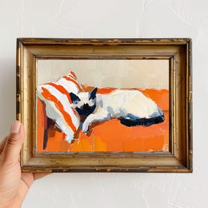 Puede incluir: Una pintura al óleo en un marco dorado representa un gato siamés descansando sobre una superficie naranja con una almohada a rayas. El gato tiene pelaje blanco con marcas negras. La pintura tiene una paleta de colores cálidos.