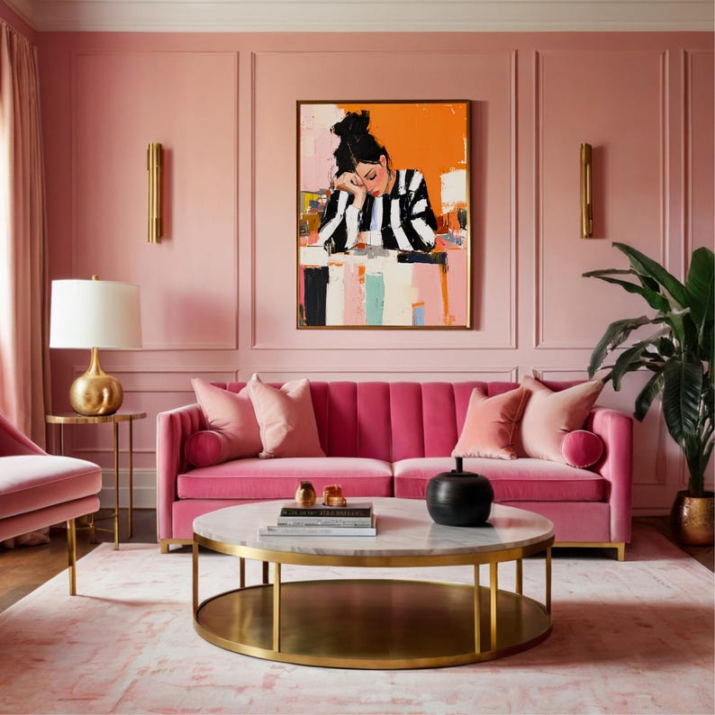 May include: A pink velvet sofa with gold legs and two pink pillows. A round coffee table with a marble top and gold legs sits in front of the sofa. A painting of a woman in a striped shirt hangs on the wall behind the sofa.