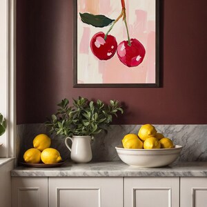 May include: A framed painting of two red cherries on a pink background with white brushstrokes. The painting is hanging on a maroon wall above a white countertop with a white pitcher and a bowl of lemons.