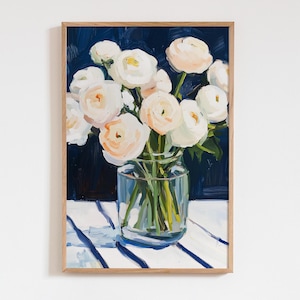 Op de afbeelding: Een ingelijst schilderij van een boeket witte en perzikbloemen in een heldere glazen vaas. De bloemen staan tegen een donkerblauwe achtergrond en een witte tafel met blauwe strepen. Het kunstwerk heeft een zachte, schilderachtige stijl.