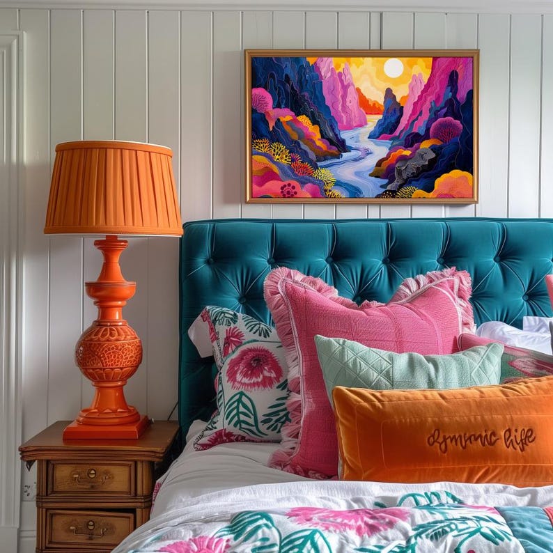 May include: A teal blue tufted headboard with a white bedspread and colorful patterned pillows. The pillows include a pink pillow with a white trim, a green pillow with a white trim, and an orange pillow with the text "dynamic life". An orange lamp with a pleated shade sits on a wooden nightstand next to the bed. A framed abstract painting of a colorful landscape hangs above the headboard.