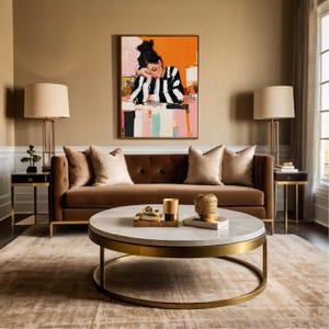 May include: A brown velvet sofa with gold legs and two throw pillows sits in a living room with a white marble coffee table with a gold base. The coffee table is decorated with a candle, a book, and a decorative bowl. A painting of a woman in a striped shirt hangs on the wall behind the sofa.