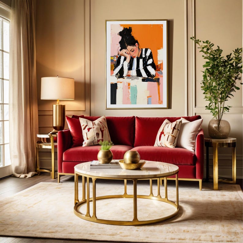 May include: A red velvet sofa with gold legs and two throw pillows sits in front of a gold coffee table with a white marble top. A painting of a woman in a striped shirt hangs on the wall behind the sofa.