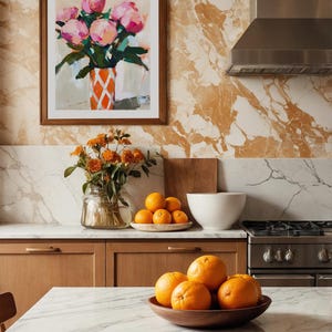 May include: A kitchen scene with a marble countertop and wooden cabinets. A framed painting of pink flowers in a vase hangs on the wall. Oranges are in a wooden bowl and on a plate.