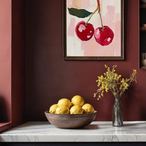 May include: A framed painting of two red cherries with green leaves on a pink and white background. The painting is hanging on a dark red wall above a bowl of yellow lemons.