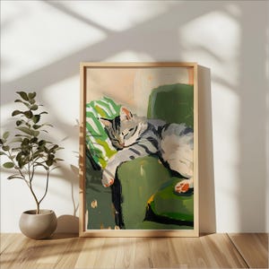 Könnte beinhalten: Gerahmtes Gemälde einer schlafenden Katze auf einem grünen Sessel. Die Katze hat grau-weiße Streifen und liegt auf einem grün-weiß gestreiften Kissen. Das Kunstwerk befindet sich in einem Holzrahmen und steht vor einer weißen Wand.