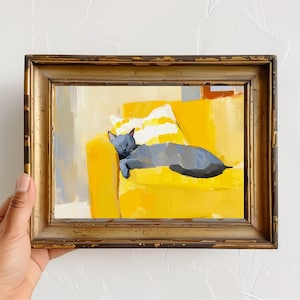 May include: An oil painting of a gray cat sleeping on a bright yellow sofa with a striped pillow. The artwork is framed in a gold-toned, ornate frame. The painting has a warm, inviting feel.