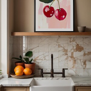 May include: A framed painting of two red cherries on a pink background. The painting is hanging on a wall above a white kitchen sink with a black faucet.