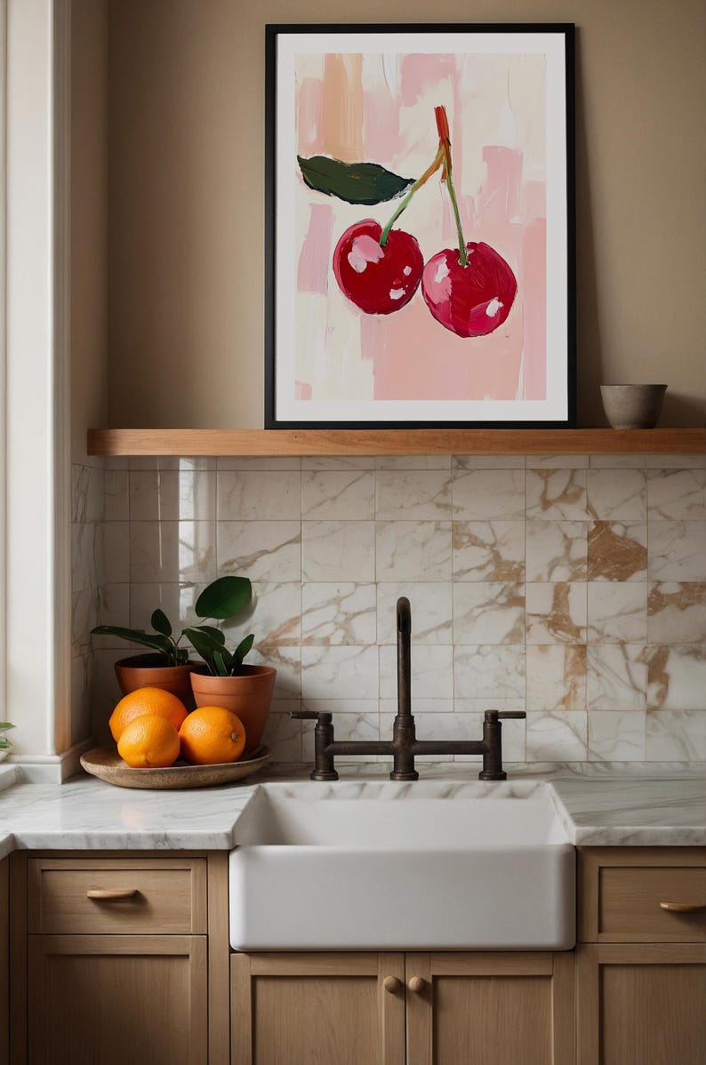 May include: A framed painting of two red cherries on a pink background. The painting is hanging on a wall above a white kitchen sink with a black faucet.