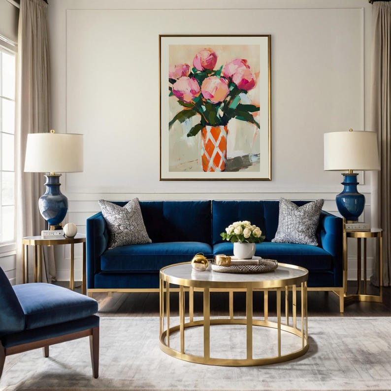 May include: A living room scene featuring a navy blue velvet sofa, a gold-framed coffee table, and two matching side tables. A large framed painting of pink flowers in an orange vase hangs above the sofa. Two navy blue lamps with white shades are on the side tables.