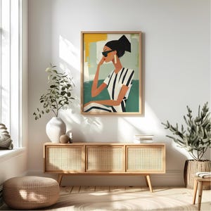 May include: A painting of a woman wearing sunglasses and a striped shirt. The woman is sitting in a chair with a green background. The painting is framed in a light wood frame.