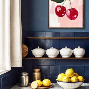 May include: A kitchen countertop with a white marble surface. A bowl filled with lemons sits on the counter, next to a small wooden tray with three lemons. A stainless steel jar and a gold metal container are also on the counter. A framed painting of two red cherries on a pink background hangs on the wall above a shelf with four white bowls.