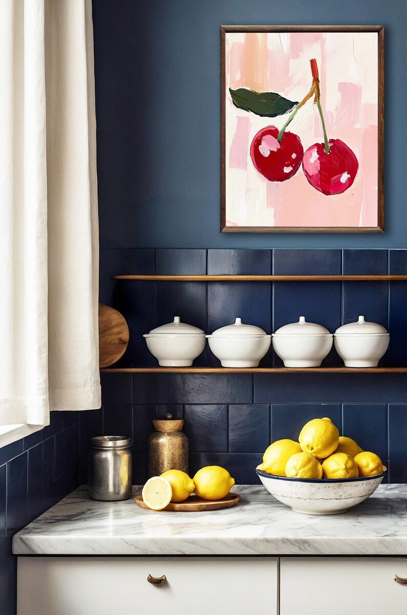 May include: A kitchen countertop with a white marble surface. A bowl filled with lemons sits on the counter, next to a small wooden tray with three lemons. A stainless steel jar and a gold metal container are also on the counter. A framed painting of two red cherries on a pink background hangs on the wall above a shelf with four white bowls.