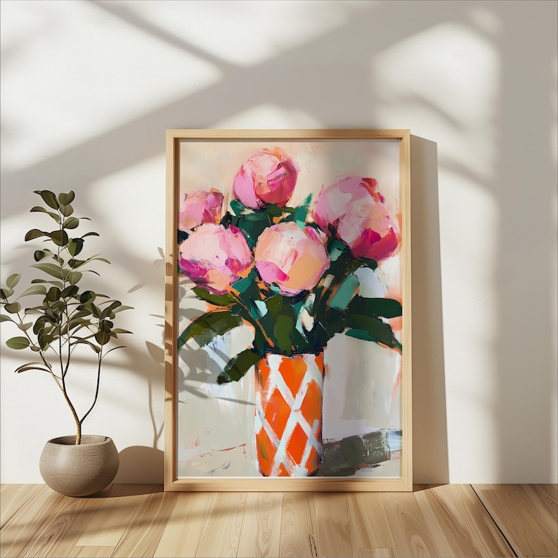 May include: A framed painting of pink peonies in a vase with an orange and white diamond pattern. The artwork has a light wood frame and is set against a neutral background. A potted plant is to the left.