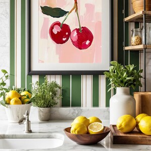 May include: A kitchen countertop with a white marble surface. A framed print of two red cherries on a pink background hangs on the wall behind a white porcelain sink. A wooden bowl filled with lemons sits on the counter next to a wooden cutting board with more lemons.