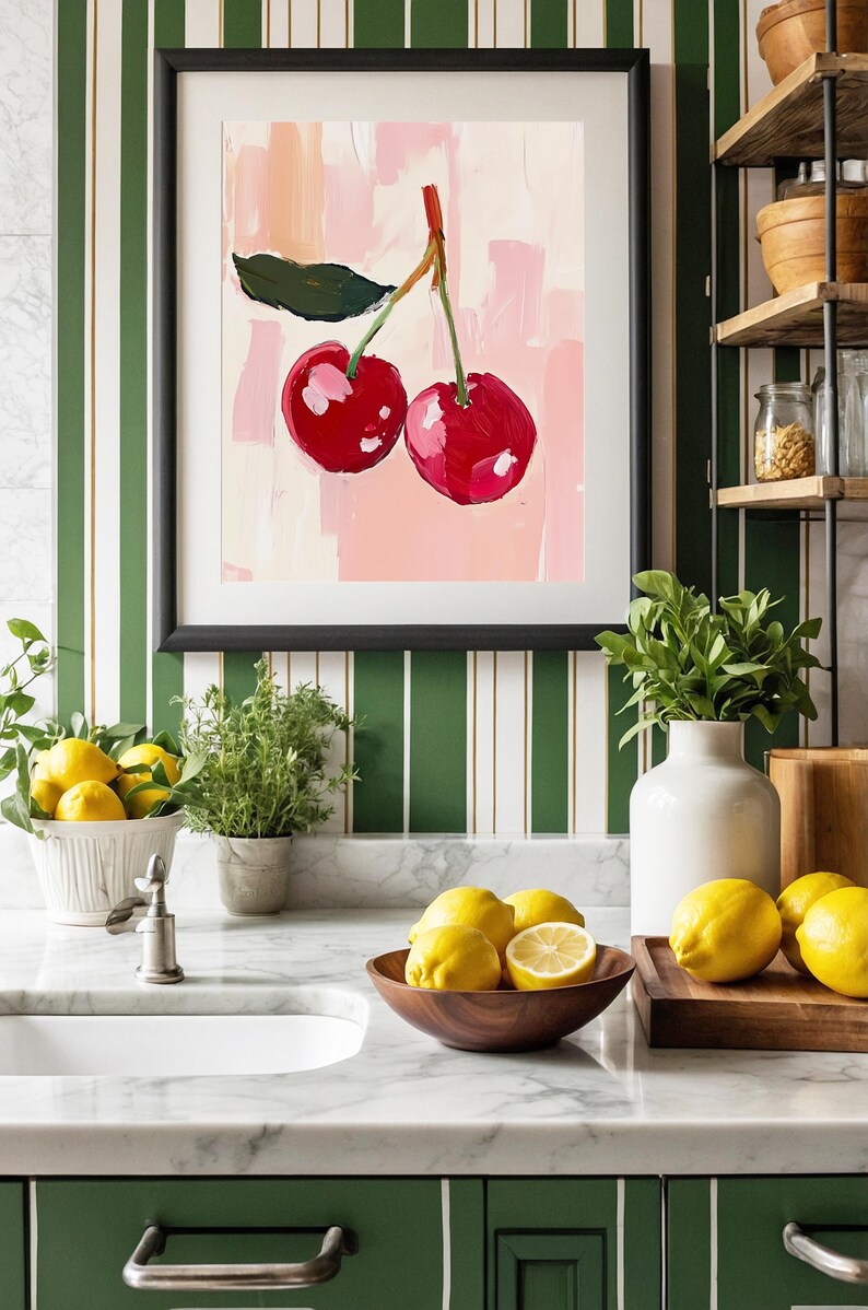 May include: A kitchen countertop with a white marble surface. A framed print of two red cherries on a pink background hangs on the wall behind a white porcelain sink. A wooden bowl filled with lemons sits on the counter next to a wooden cutting board with more lemons.