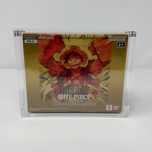 May include: A sealed box of "ONE PIECE CARD GAME" trading cards. The box is gold with a vibrant illustration of a character. Text includes "THE BEST ONE PIECE CARD GAME" and "10 CARDS PER PACK / 20 PACKS PER BOX". The box is in a clear acrylic case.