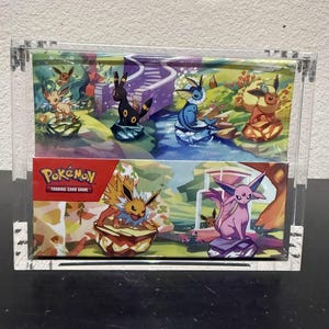May include: A clear acrylic display case holding two Pokémon Trading Card Game boxes. The top box features a colorful illustration of several Pokémon characters near a winding path. The bottom box has the Pokémon Trading Card Game logo.