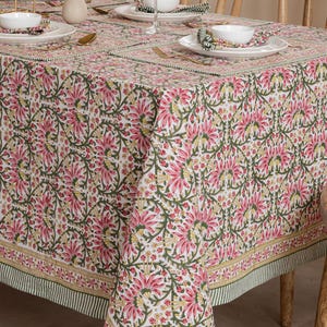 May include: A floral tablecloth with a repeating pattern of pink flowers, green leaves, and yellow accents on a white background. The tablecloth is set on a wooden table with place settings, including white plates, bowls, and gold-colored cutlery.