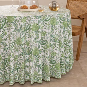 May include: A round tablecloth featuring a white backdrop and a repeating green floral design. The table is set with a glass dome, a teapot, and a glass of wine. A wooden chair is in the background.