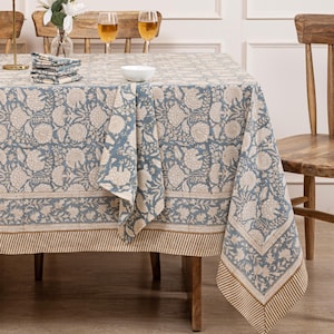 May include: A blue and white floral tablecloth with a brown border. The tablecloth is laid on a wooden table with two chairs and two wine glasses.