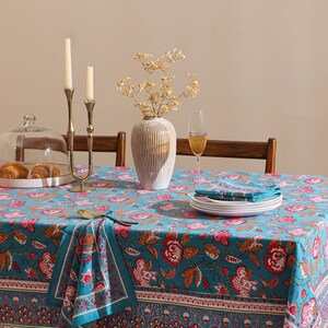 May include: A rectangular tablecloth featuring a vibrant turquoise background with a repeating floral pattern in shades of red, orange, and white. The fabric includes a decorative border with intricate botanical motifs and matching folded napkins.