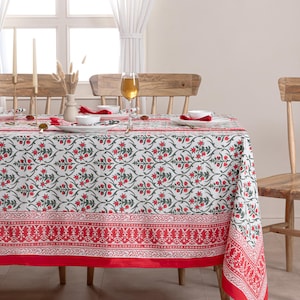 Könnte beinhalten: Eine weiße Tischdecke mit rotem Blumenmuster und rotem Rand liegt auf einem Holztisch. Der Tisch ist mit weißen Tellern, roten Servietten und Weingläsern gedeckt. Zwei Holzstühle sind zu sehen.