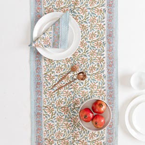 May include: A white floral table runner with a blue striped border, set with white plates, a folded napkin, and copper-colored utensils. A small bowl holds three red pomegranates, creating a vibrant dining scene.